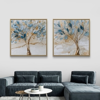 סט זוג תמונות קנבס מרובעות עם עצים אמנותיים בגווני כחול ותכלת ונגיעות זהב על רקע שמנתי, תלויות מעל ספה מודרנית.