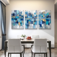 תמונת זכוכית מחולקת לשלושה חלקים בעיצוב אבסטרקטי בגווני כחול, טורקיז וזהב תלויה מעל שולחן אוכל מודרני.