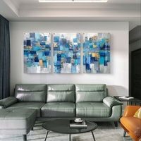 תמונת זכוכית מחולקת לשלושה חלקים בעיצוב אבסטרקטי בגווני כחול, טורקיז וזהב תלויה מעל שולחן אוכל מודרני.