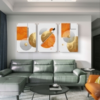 תמונת זכוכית מחולקת לשלושה חלקים בעיצוב גיאומטרי בגווני כתום וזהב תלויה מעל ספה מודרנית בסלון.