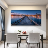 תמונת זכוכית בהדפסה חלקה של מזח עץ המוביל אל עיר מוארת בלילה עם השתקפויות על המים, בעיצוב מודרני ויוקרתי.