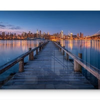 תמונת זכוכית בהדפסה חלקה של מזח עץ המוביל אל עיר מוארת בלילה עם השתקפויות על המים, בעיצוב מודרני ויוקרתי.