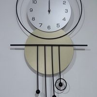 שעון קיר מעוצב בסגנון מודרני עם צורות גאומטריות, מסגרת עגולה, מטוטלות דקורטיביות וגימור אלגנטי בגווני שחור, לבן וזהב, תלוי על קיר בהיר.