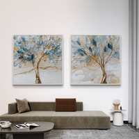סט זוג תמונות קנבס מרובעות עם עצים אמנותיים בגווני כחול ותכלת ונגיעות זהב על רקע שמנתי, תלויות מעל ספה מודרנית.