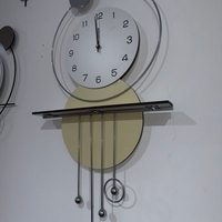 שעון קיר מעוצב בסגנון מודרני עם צורות גיאומטריות, מסגרת עגולה, מטוטלות דקורטיביות וגימור אלגנטי בגווני זהב, אפור ולבן, תלוי על קיר בעיצוב מינימליסטי.