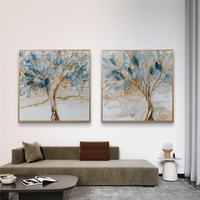 סט זוג תמונות קנבס מרובעות עם עצים אמנותיים בגווני כחול ותכלת ונגיעות זהב על רקע שמנתי, תלויות מעל ספה מודרנית.