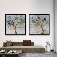 סט זוג תמונות קנבס מרובעות עם עצים אמנותיים בגווני כחול ותכלת ונגיעות זהב על רקע שמנתי, תלויות מעל ספה מודרנית.