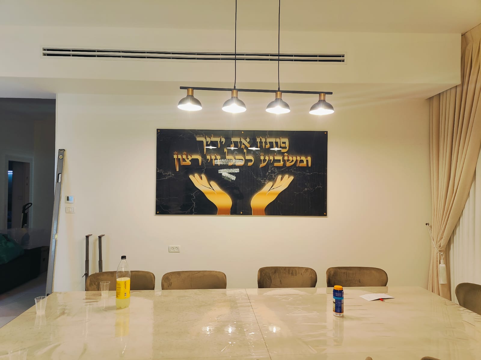 תמונת זכוכית יוקרתית עם טקסט מעורר השראה תלויה מעל פינת אוכל מודרנית. שילוב מושלם של עיצוב אלגנטי, שולחן שיש לבן וכיסאות מרופדים בגוון בהיר, המשדרים ניקיון וסטייל עכשווי.