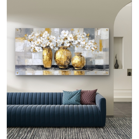 תמונת זכוכית לרוחב עם שלושה אגרטלים מוזהבים מלאים בפרחים לבנים על רקע מופשט בגווני אפור וזהב, תלויה מעל ספה מודרנית כחולה.