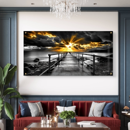 תמונת זכוכית אומנותית לרוחב המציגה שביל עם מעקה המוביל לים, בשחור־לבן עם שקיעה זהובה דרמטית, תלויה מעל ספה בסלון אלגנטי.