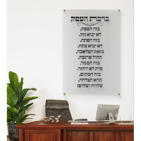 תמונת זכוכית שקופה עם כיתוב ברכת העסק בצבע שחור תלויה על קיר לבן במשרד יוקרתי עם שולחן עץ כהה, כסא מנהלים וצמח ירוק בצד.