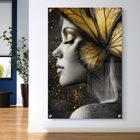 תמונת זכוכית של אישה בפרופיל עם כנפי פרפר זהובות המשתלבות בפניה, על רקע שחור עם נצנוצים זהובים. תמונה תלויה על קיר לבן בחלל מודרני.