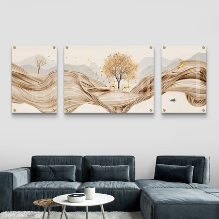 שלוש תמונות זכוכית מחולקות עם עיצוב קווים זורמים בגווני זהב, עץ במרכז, צבאים ועיצוב טבע פסטורלי, תלויות מעל ספה בהירה בקיר אפור בהיר.          Ask ChatGPT