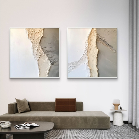 זוג תמונות קנבס מרובעות בגוונים רגועים של לבן, שמנת ואפור עם מרקם תלת־ממדי, תלויות על קיר לבן מעל ספה מודרנית בגוון חום־אפרפר.