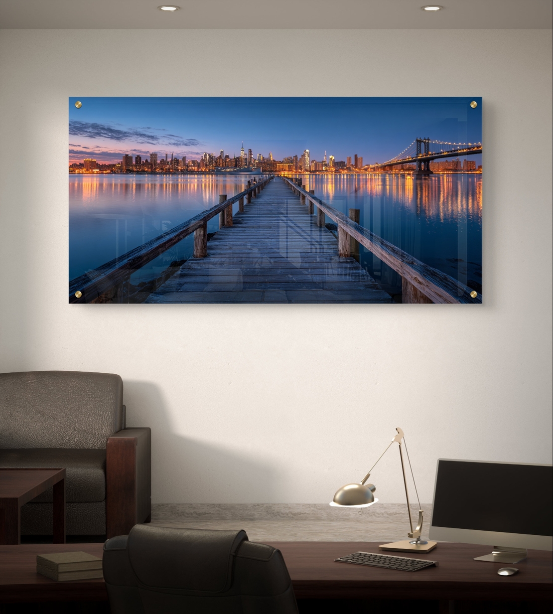 תמונת זכוכית בהדפסה חלקה של מזח עץ המוביל אל עיר מוארת בלילה עם השתקפויות על המים, בעיצוב מודרני ויוקרתי.