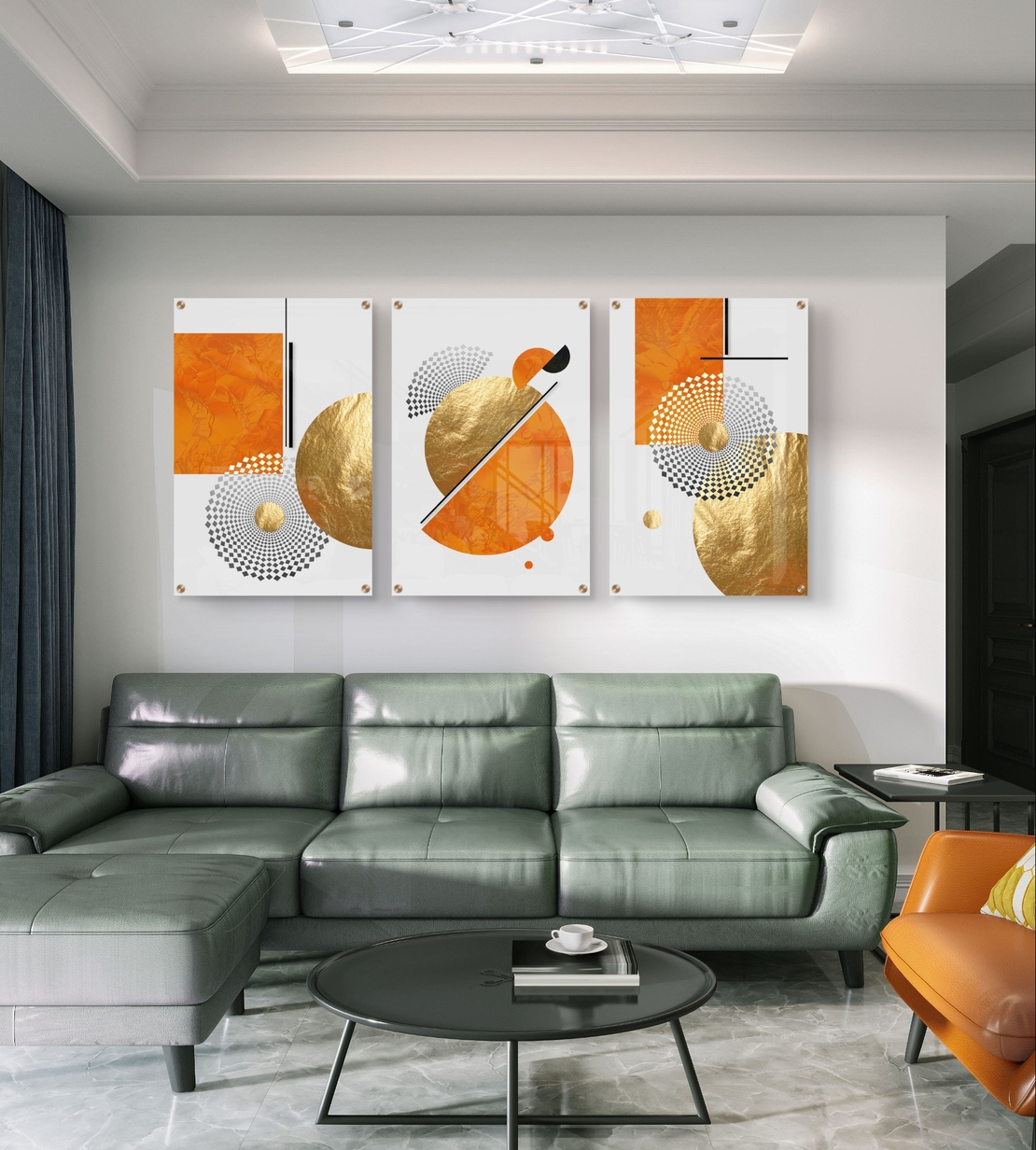 תמונת זכוכית מחולקת לשלושה חלקים בעיצוב גיאומטרי בגווני כתום וזהב תלויה מעל ספה מודרנית בסלון.