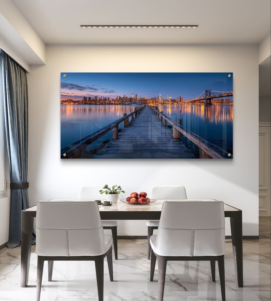 תמונת זכוכית בהדפסה חלקה של מזח עץ המוביל אל עיר מוארת בלילה עם השתקפויות על המים, בעיצוב מודרני ויוקרתי.