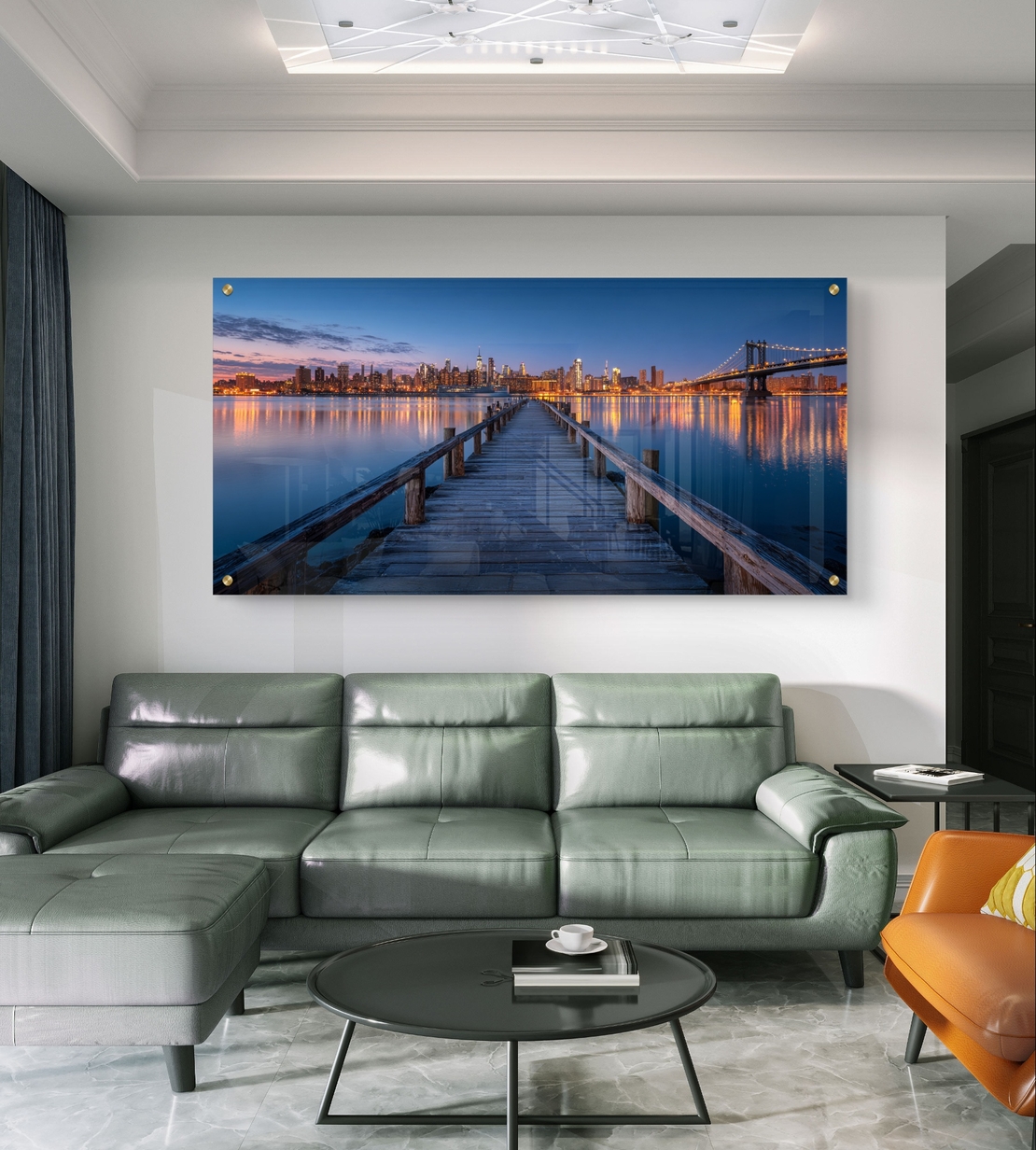 תמונת זכוכית בהדפסה חלקה של מזח עץ המוביל אל עיר מוארת בלילה עם השתקפויות על המים, בעיצוב מודרני ויוקרתי.